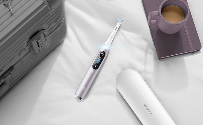 Oral-B iO toothbrush in lavender, with travel case, suitcase, and coffee mug on a white surface.