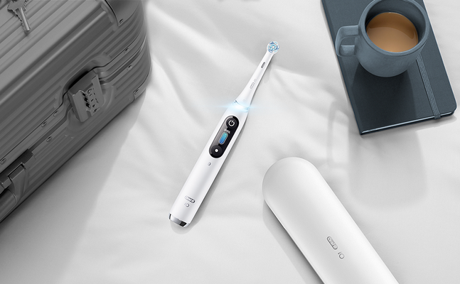 Oral-B iO toothbrush displayed with its travel case, a suitcase, and a cup of coffee on a notebook.