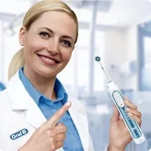 A smiling woman in a lab coat points to an electric toothbrush she is holding with the text Oral-B on it.