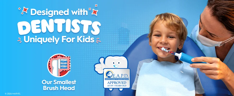 A dentist helps a young child brush with a blue electric toothbrush featuring a Spider-Man design. Text: “Designed with Dentists, Uniquely for Kids” and “Our Smallest Brush Head.”