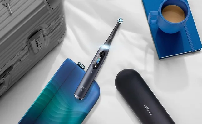 Oral-B iO toothbrush displayed with travel case, charging case, suitcase, notebook, and coffee cup.