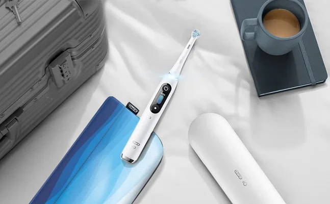 Oral-B iO toothbrush displayed with travel case, charger, suitcase, and coffee cup on a white surface.