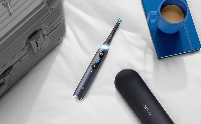 Oral-B iO toothbrush with charging display, travel case, suitcase, notebook and coffee mug on a white surface.