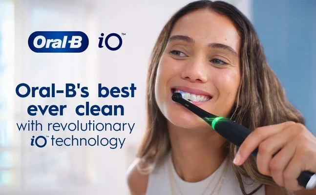 A woman is smiling while using a black electric toothbrush with a glowing green indicator, with the text Oral-Bs best ever clean with revolutionary iO technology displayed in blue and black.